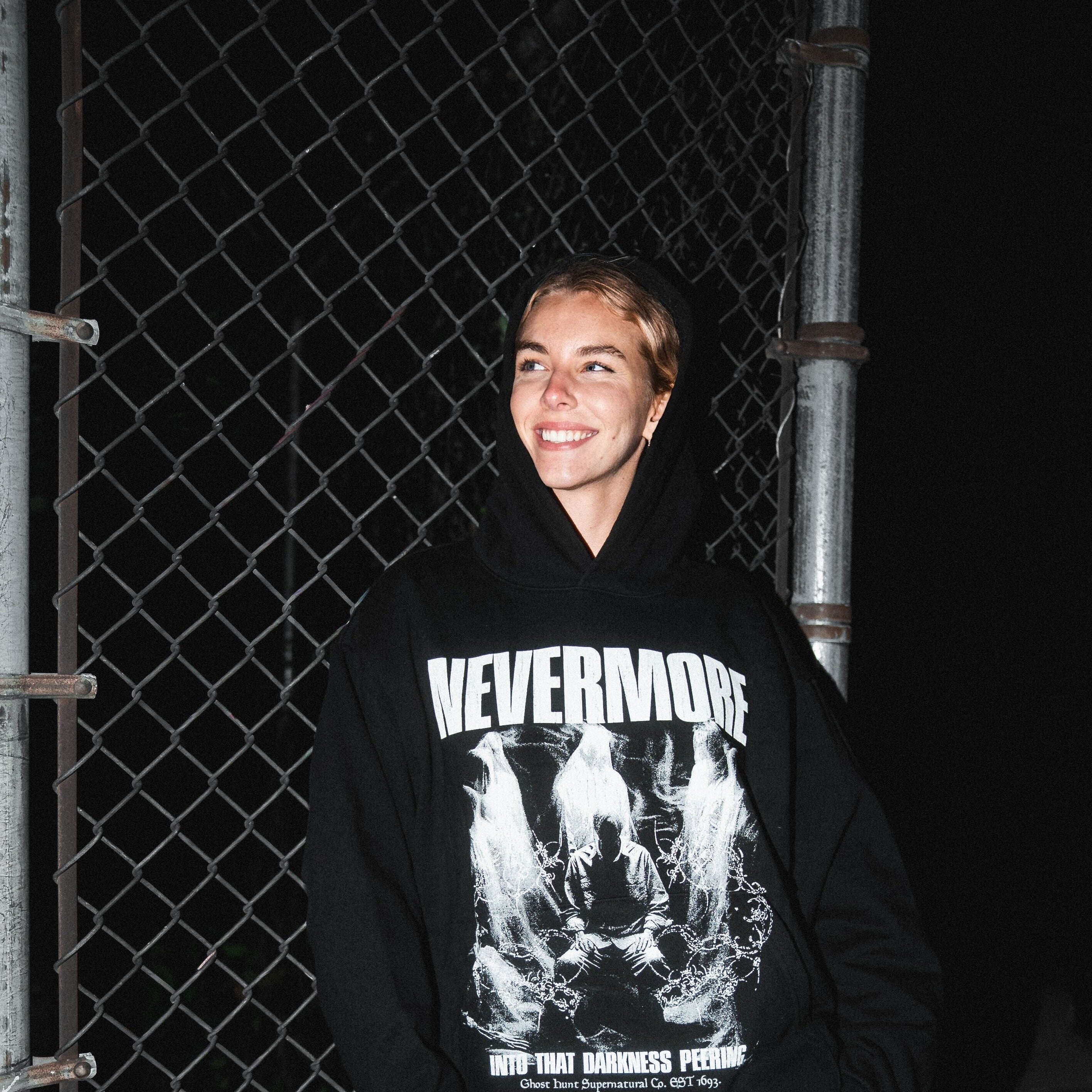 Person wearing a black hoodie with 'Nevermore' design in front of a chain-link fence.