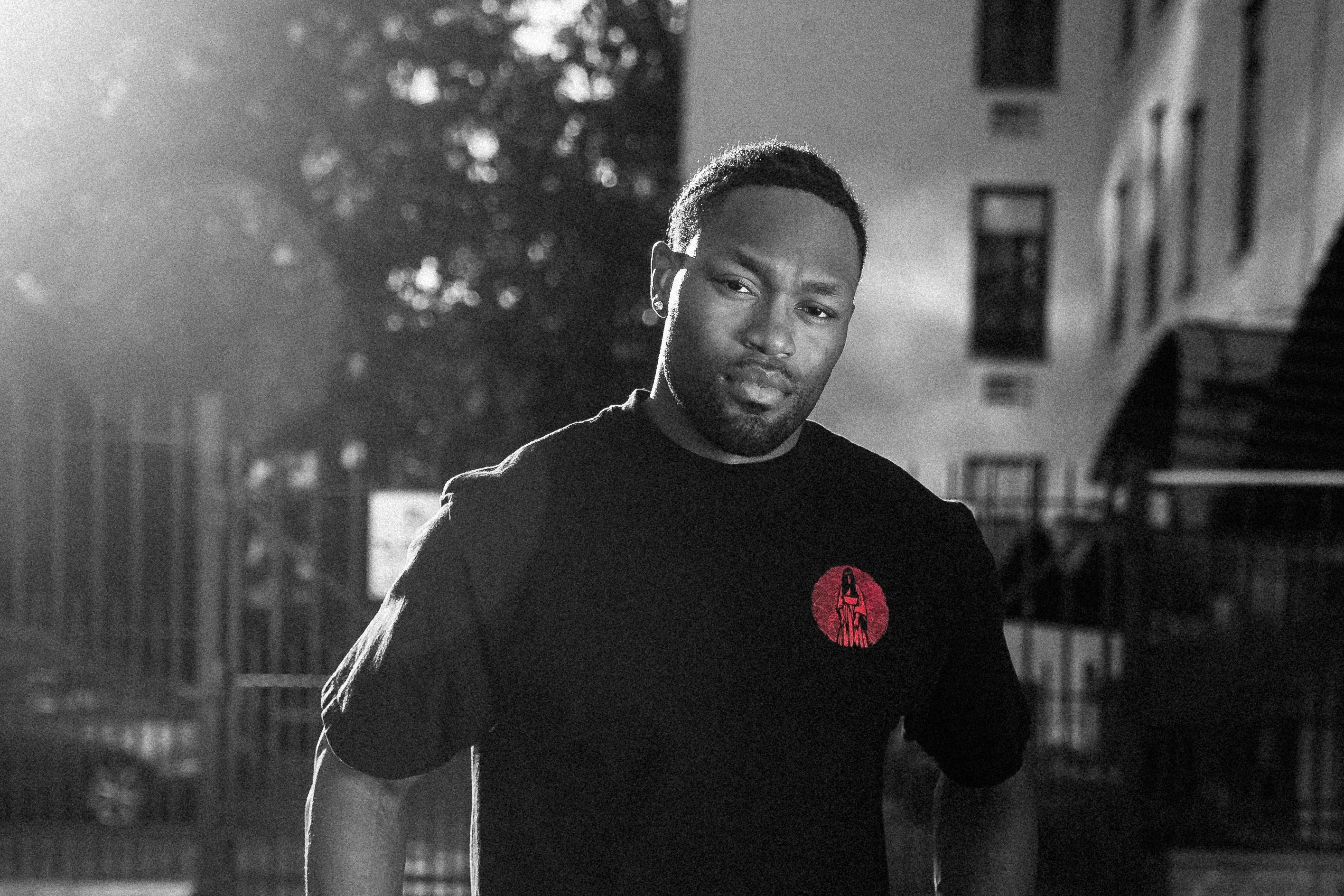 A man wearing a black crewneck t-shirt with a small red circular graphic on the left chest, paired with dark jeans, standing outdoors in a city setting with strong sunlight and shadows.