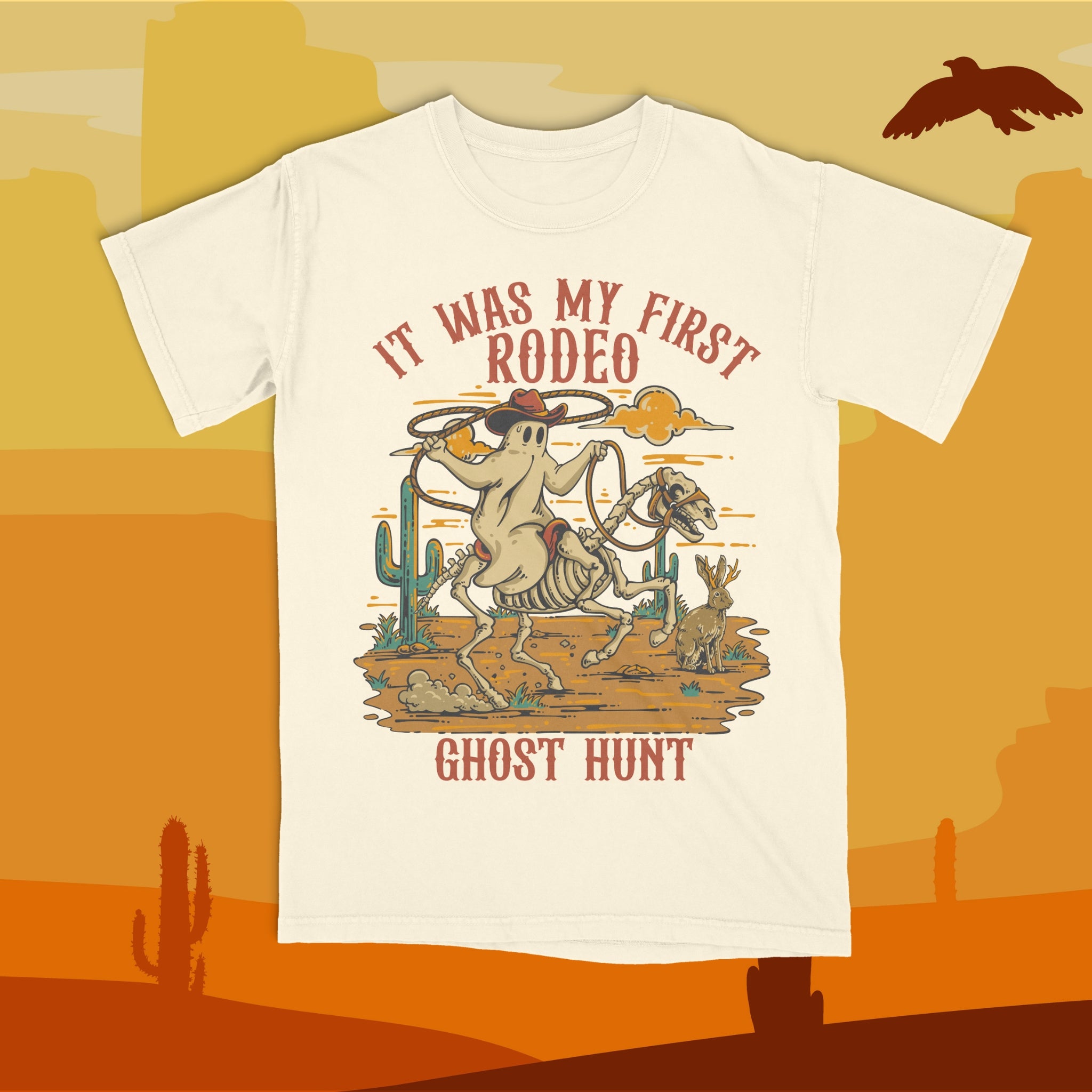 White t-shirt with a graphic design of a skeleton riding a bucking bronco with text 'It Was My First Rodeo Ghost Hunt' on an orange desert background.